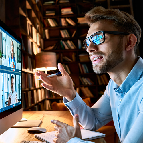 Man in blue shirt  and black glasses, sat a desk on an online meeting, holding a men, talking in an home office with books in the background.