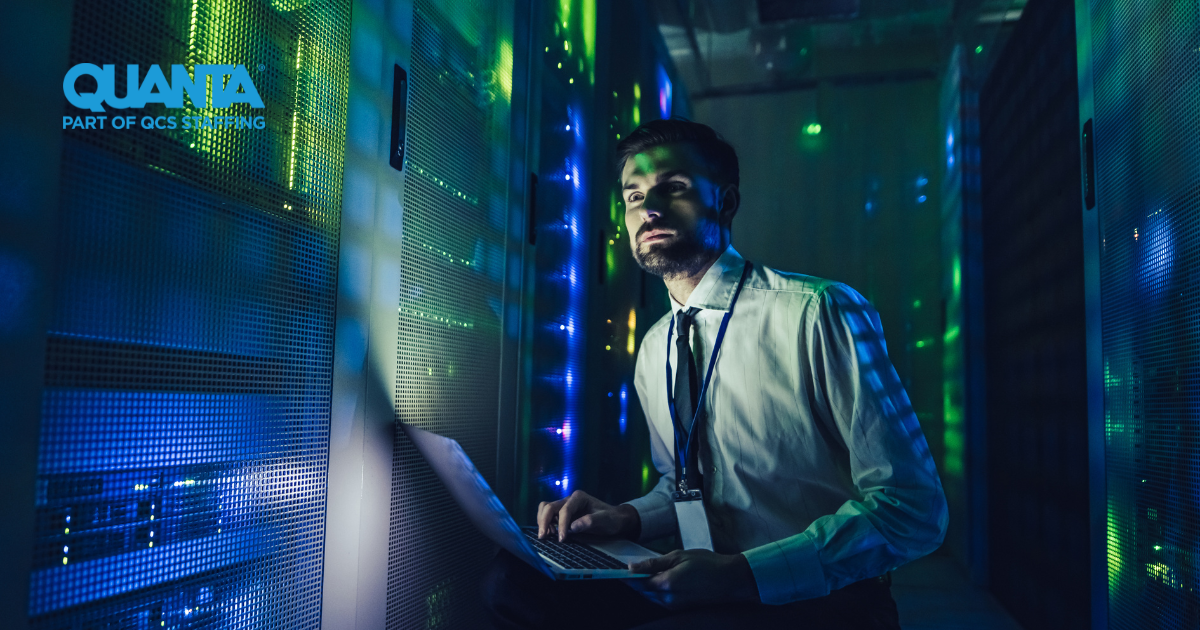 man in shirt and tie typing on a laptop and looking at data centre computers