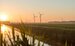 5 wind turbines in a field next to a stream with a sunset behind them