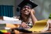 A young graduate wearing a graduation gap and glasses, sat in front of a pile of booka.