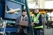 Two men, one in a high visibility jacket and a white hard hat holding a planner, the other in a suit holding a laptop.