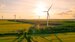 AERIAL, LENS FLARE: Colorful sunset sky above farmland and spinning windmills. Picturesque English countryside with tall modern power generation