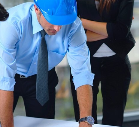 A man in a blue shirt, tie, and hard hat looking at something on a table