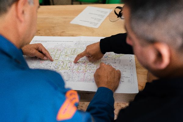 Two men with their back to the camera, they are both pointing at a blueprint on a wooden table.