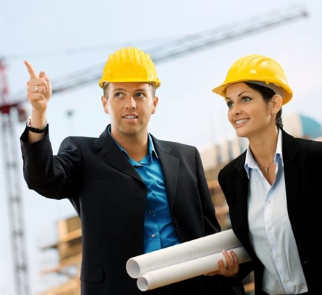 a man and a woman in suits and hardhats, the man is pointing up while holding some rolled up paper
