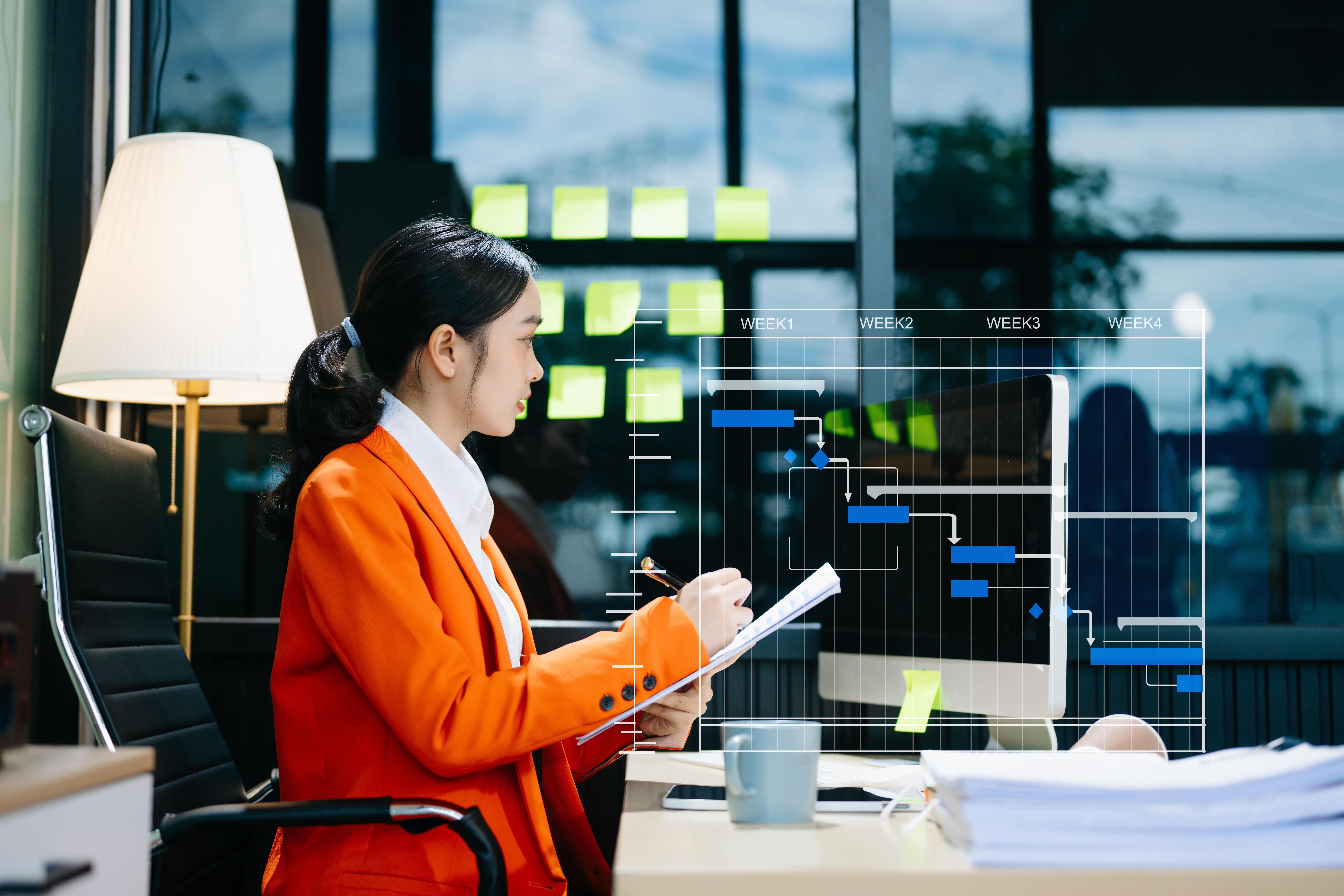 A lady in an office chair in an orange suit jacket and shite shirt with a virtual project management screen central to the image.