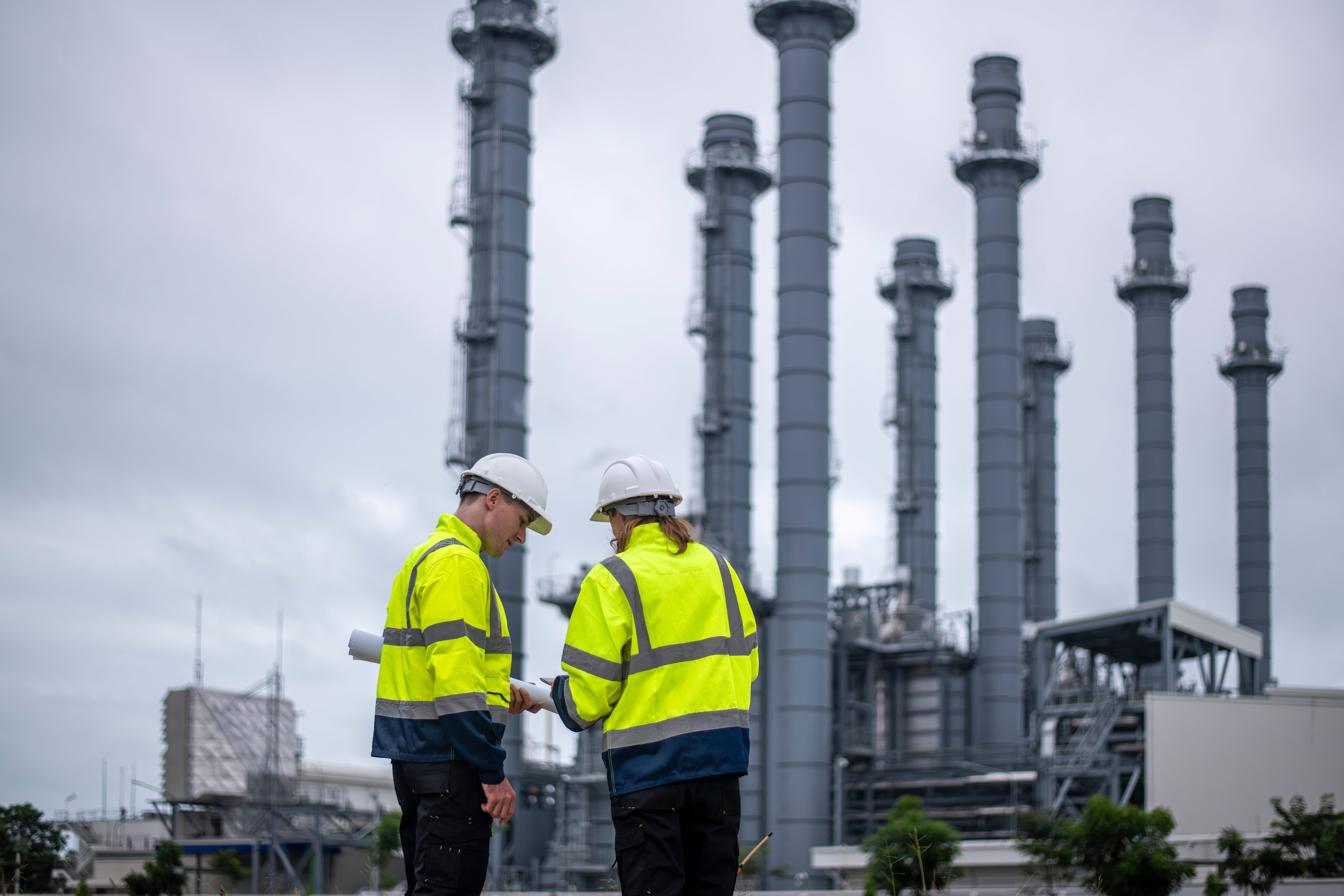 Engineers wearing safety gear, including hard hats examining survey a large blueprint tablet standing industrial facility gas or oil refinery