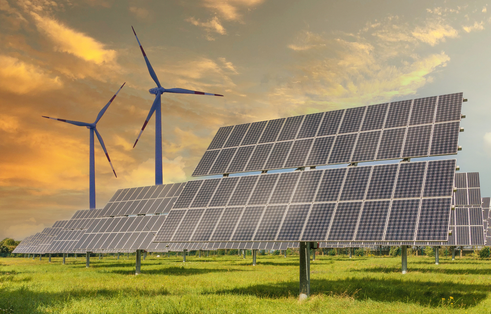 Wind turbines and solar panels on a green field with a sunset sky as the backdrop.