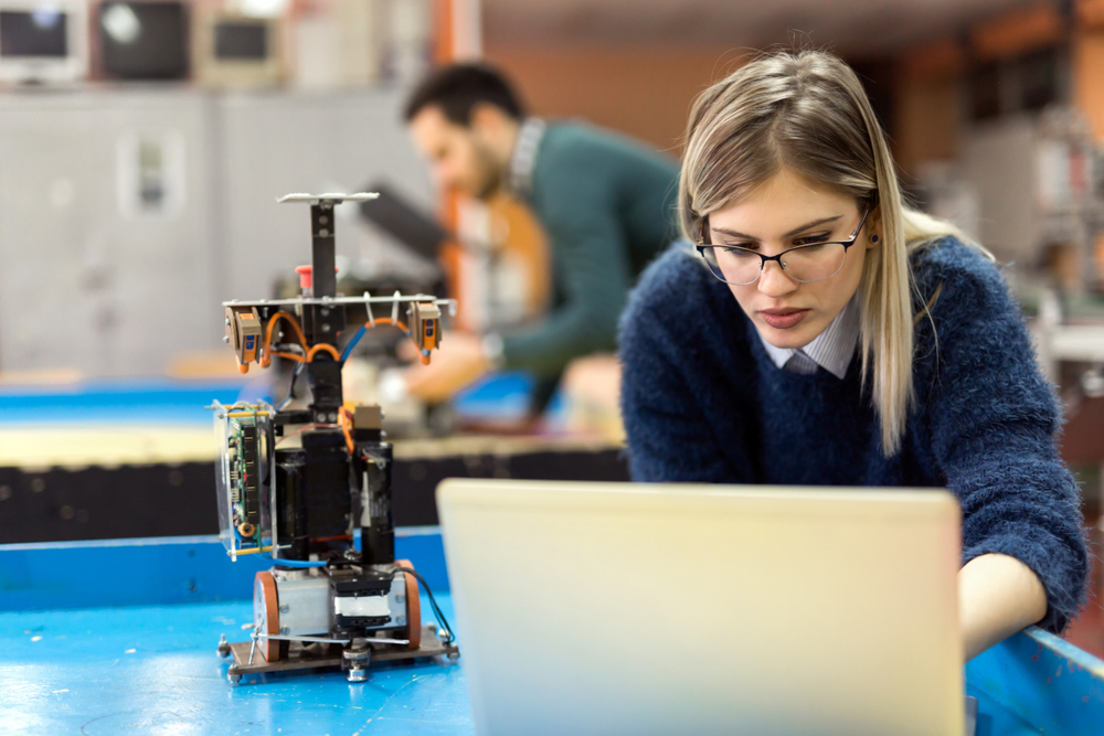 A blond lady with glasses sat in a workshop in front of a laptop