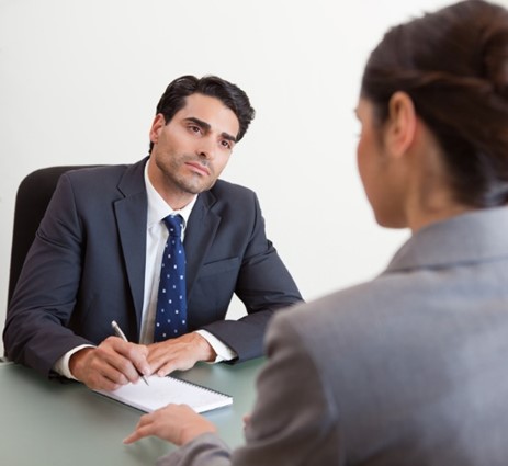 a man in a suit and tie holding a pen and paper, interviewing a woman with her back to the camera.