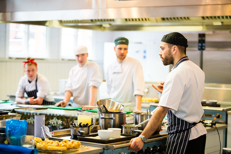 man in chef whites and apron explaining something to three people people in chef whites