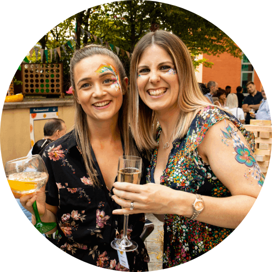 two employees smiling and enjoying cocktails at a work social event outdoors with fun face glitter, it appears to be summertime