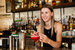 Young woman serving bright cocktail on a bar with fully stocked shelf of spirits as a backdrop