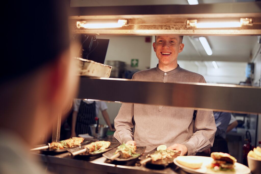 A smiling man in a grey shirt in a restaurant kitchen stood in front of four plates of food.