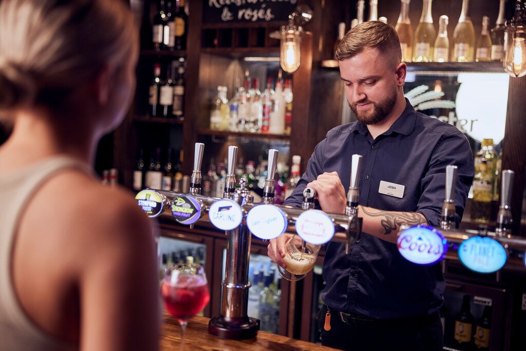 A man standing behind a bar, pulling a pint of beer for a woman.