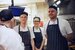 4 people stood in a kitchen, one with their back to the camera, each are wearing striped aprons and chef hats.