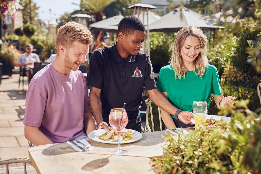 3 people sat in a restaurant garden, the waiter stands in the middle of the two people at the table.  