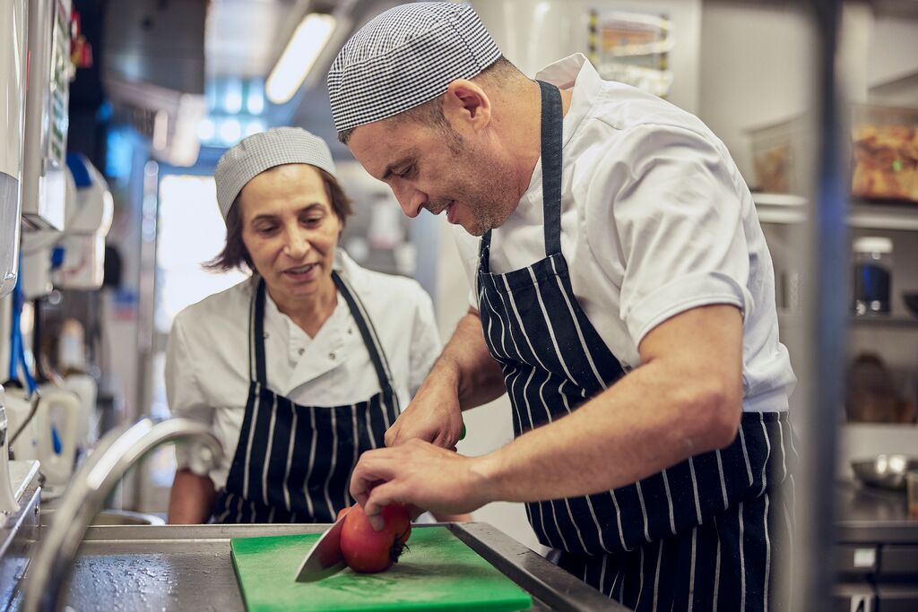 sous chef standing over a kitchen apprentice watching them prep food