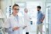 Optician wearing glasses and lab coat holding iPad standing in store with two people in the background talking