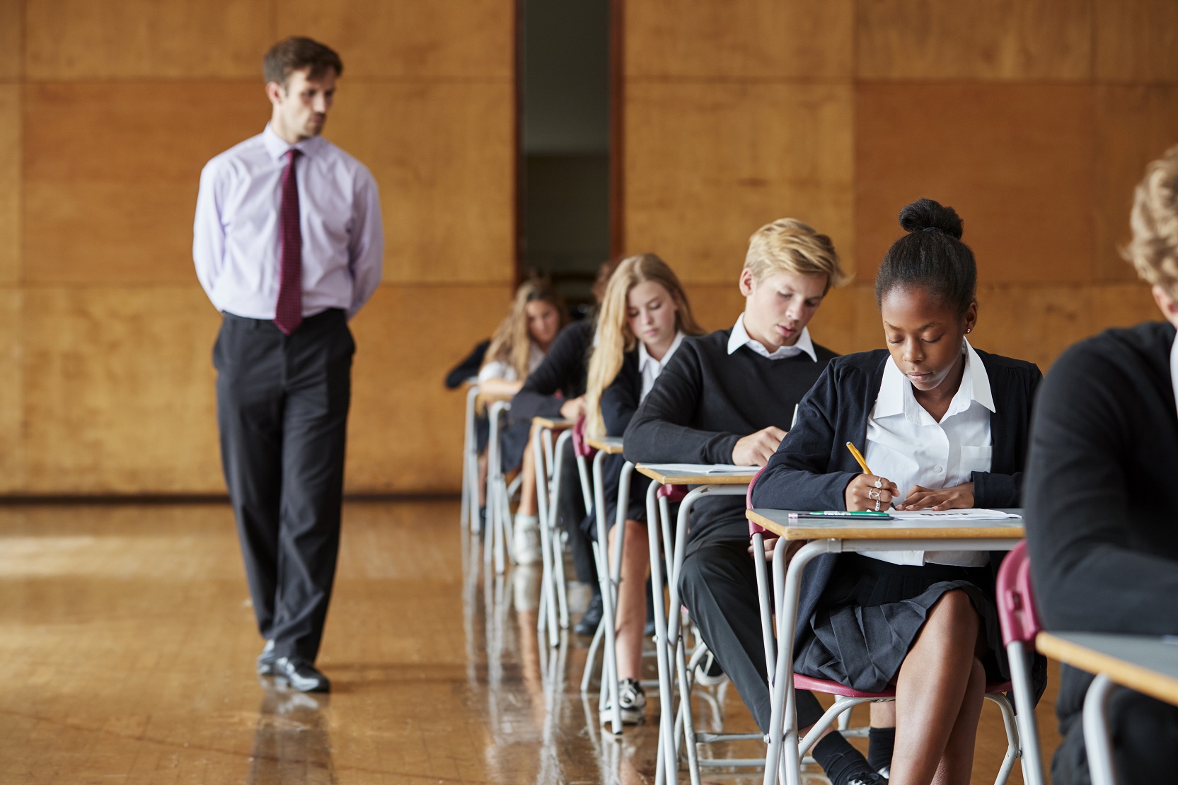 Teacher in shirt and tie supervising students at desks taking exams in a school hall