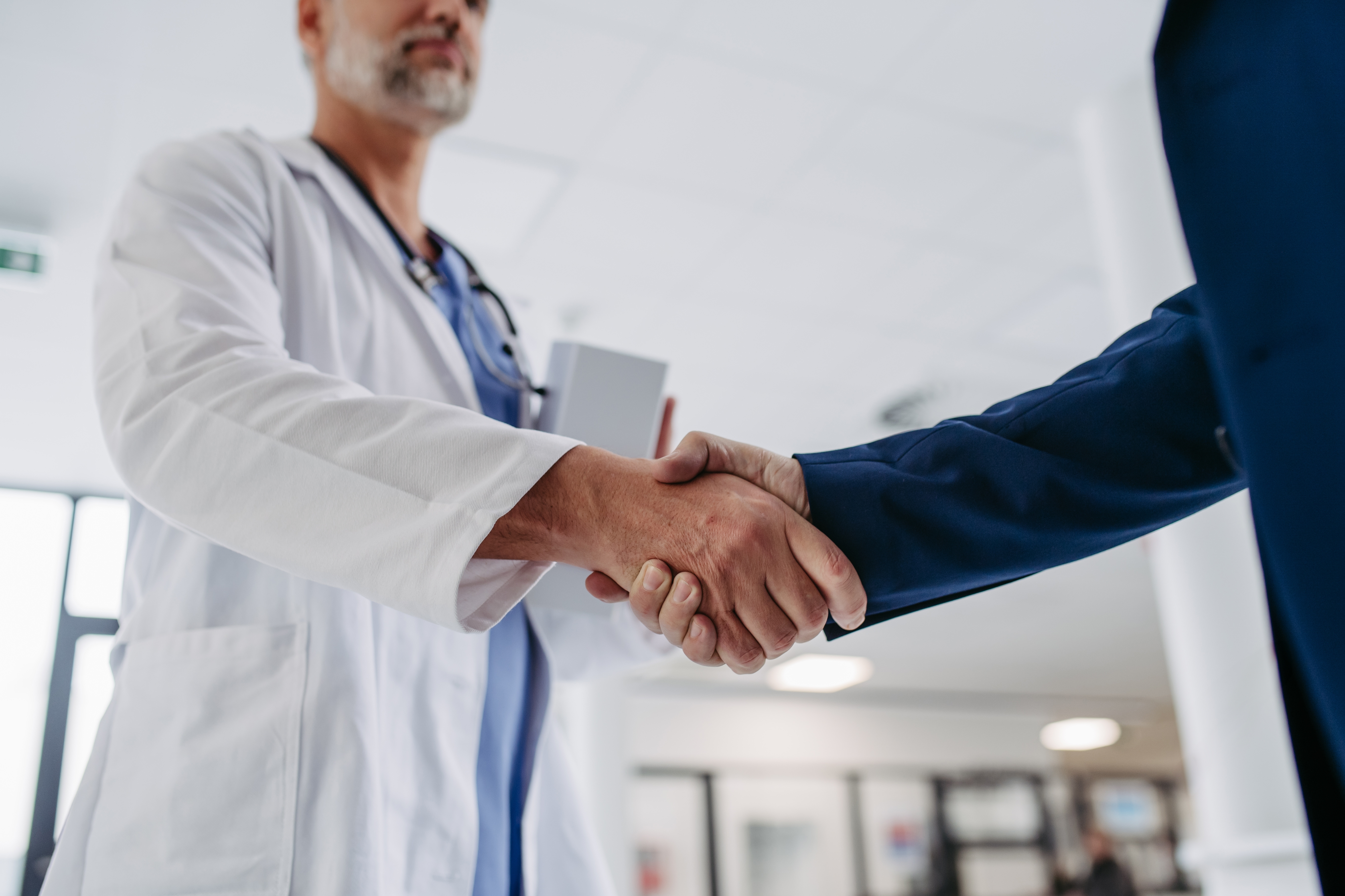 Close up of pharmaceutical sales representative presenting new medication to doctor in medical building, shaking hands.