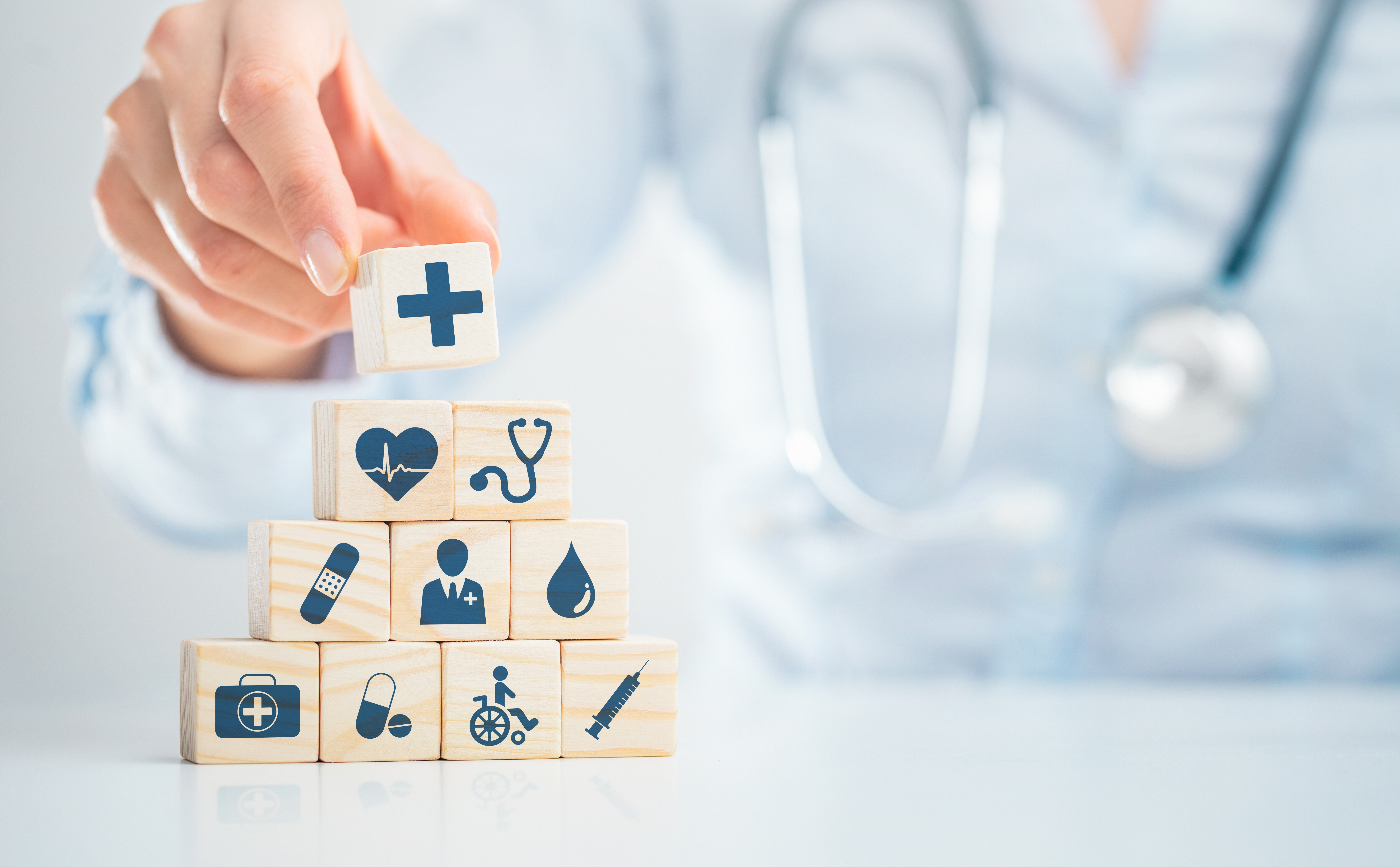 A hand placing a wooden blog on top of a pyramid shaped stack of 9 other blogs, each have symbols relating to healthcare on them in dark blue.