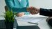 a resume sits on a desk next to a plant and a laptop. Two people are shaking hands over it