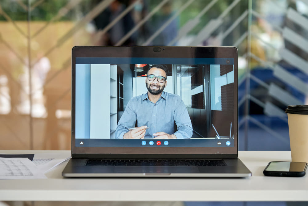 Laptop computer at office workplace table with smiling indian businessman executive leader, coach on pc screen having video chat, conference call, virtual meeting or giving training on videocall