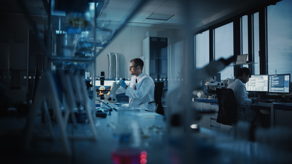 man in white lab coat sitting in a laboratory with a microscope