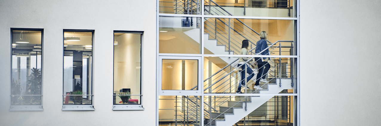 a modern office space with multiple windows and people walking on the staircase visible through glass 