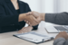 Two people shaking hands over a desk with a clipboard and laptop on a grey table 