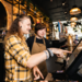 An apprentice being shown how to operate the till behind a Greene King bar by a senior member of staff