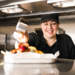 A smiling female Greene King chef pouring sauce onto a dish in the kitchen