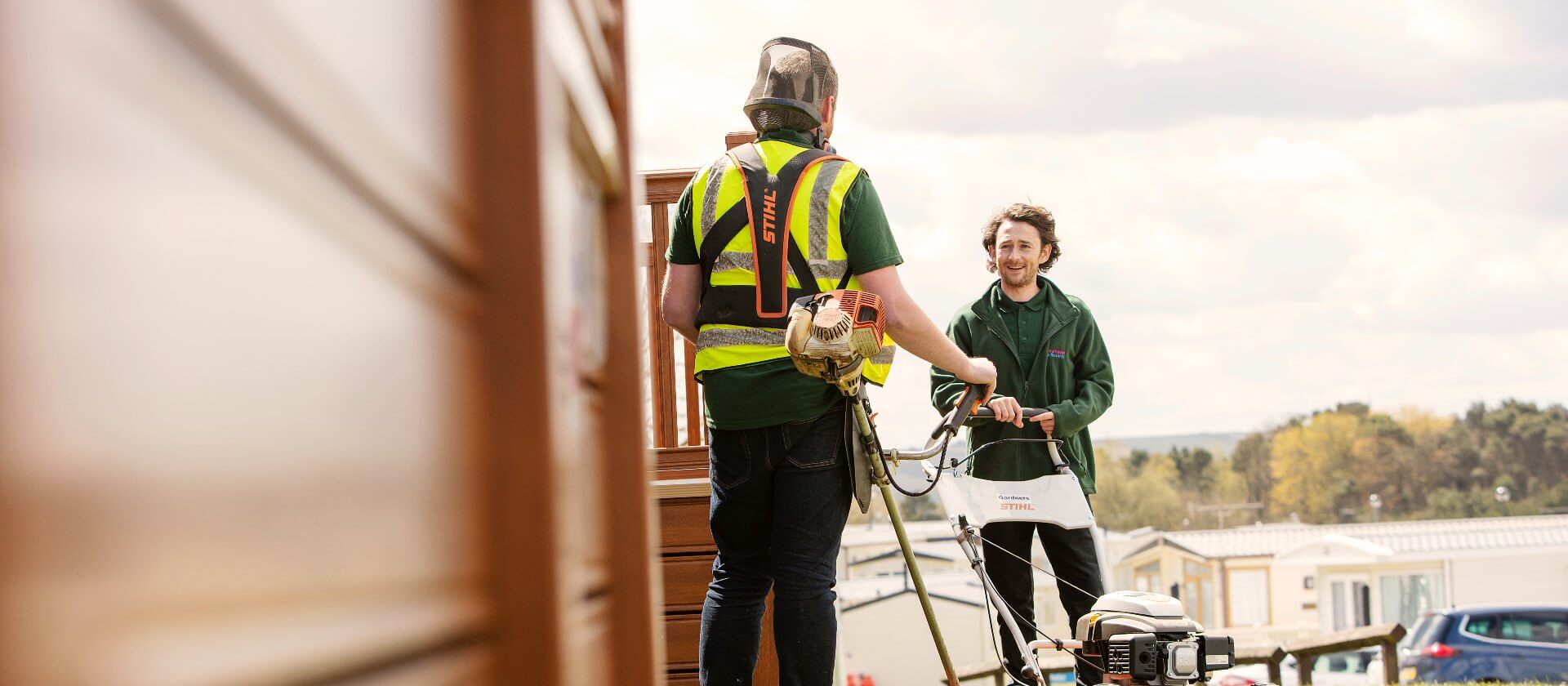 Image of man mowing the lawn and speaking with another man dressed in green clothing.