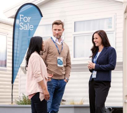 An image of three adults having a conversation in front of a holiday home and for sale sign.