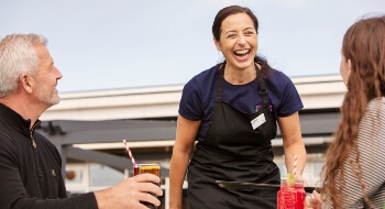 Waitress in apron is laughing while she serves a man and a woman drinks outside