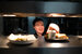 A person front and centre image in a restaurant kitchen reaching towards two plates of food.