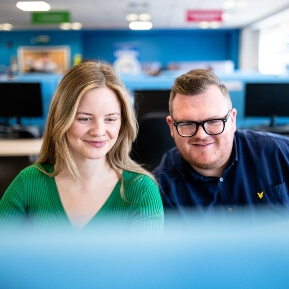 a man and a woman, sat smiling as they look at a computer screen in front of them