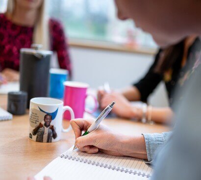 Am image of a woman writing in a notebook with a pen in an office setting