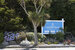 The Lizard Point Holiday Park sign is blue and sits behind a tree with some greenery and flowers around it.