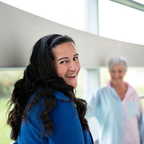 a woman looks over her shoulder smiling, as another woman stand in the background smiling