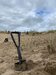 Some sand dunes on a beach with a shovel front and centre of the image, people are in the background, cleaning up the beach