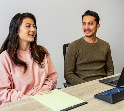 girl in pink jumper and boy in green top sat at a desk looking at each other and smiling