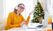 Cheerful woman in glasses and headphones smiling and speaking with online colleague on Christmas day at home