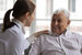 Caring caregiver in white coat cares for grey-haired elderly man in nursing home, listening, providing help during visit at home. Homecare eldercare caregiving concept