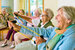 Large group of happy enthusiastic elderly ladies being led by a caregiver, exercising in a gym sitting in chairs doing stretching exercises with rubber bands