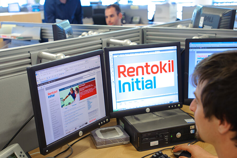 A worker in a cubicle, focused on a computer displaying the Rentokil Initial logo and website on multiple monitors.
