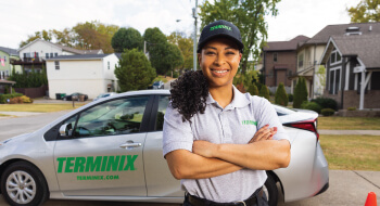 A smiling Terminix employee in a grey shirt and cap stands with her arms crossed in front of a Terminix car.
