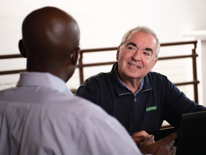 Two men are sitting at a table, talking. A man wearing a blue sweater with the Terminix logo is smiling at his conversation partner.