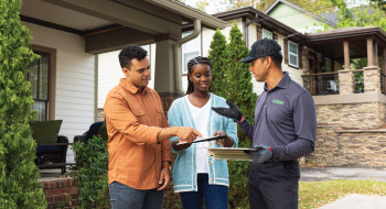 A Terminix technician wearing a gray shirt and black cap stands in front of a house and talks to a smiling couple.